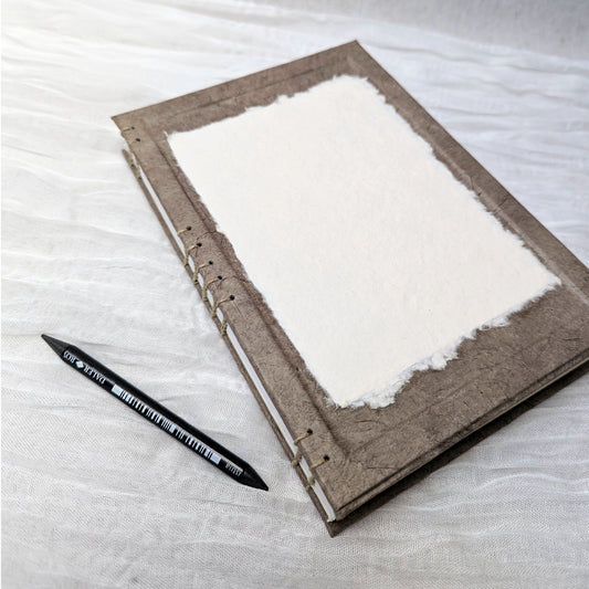 Brown art journal with white pages on a light gray surface, accompanied by a black pen.