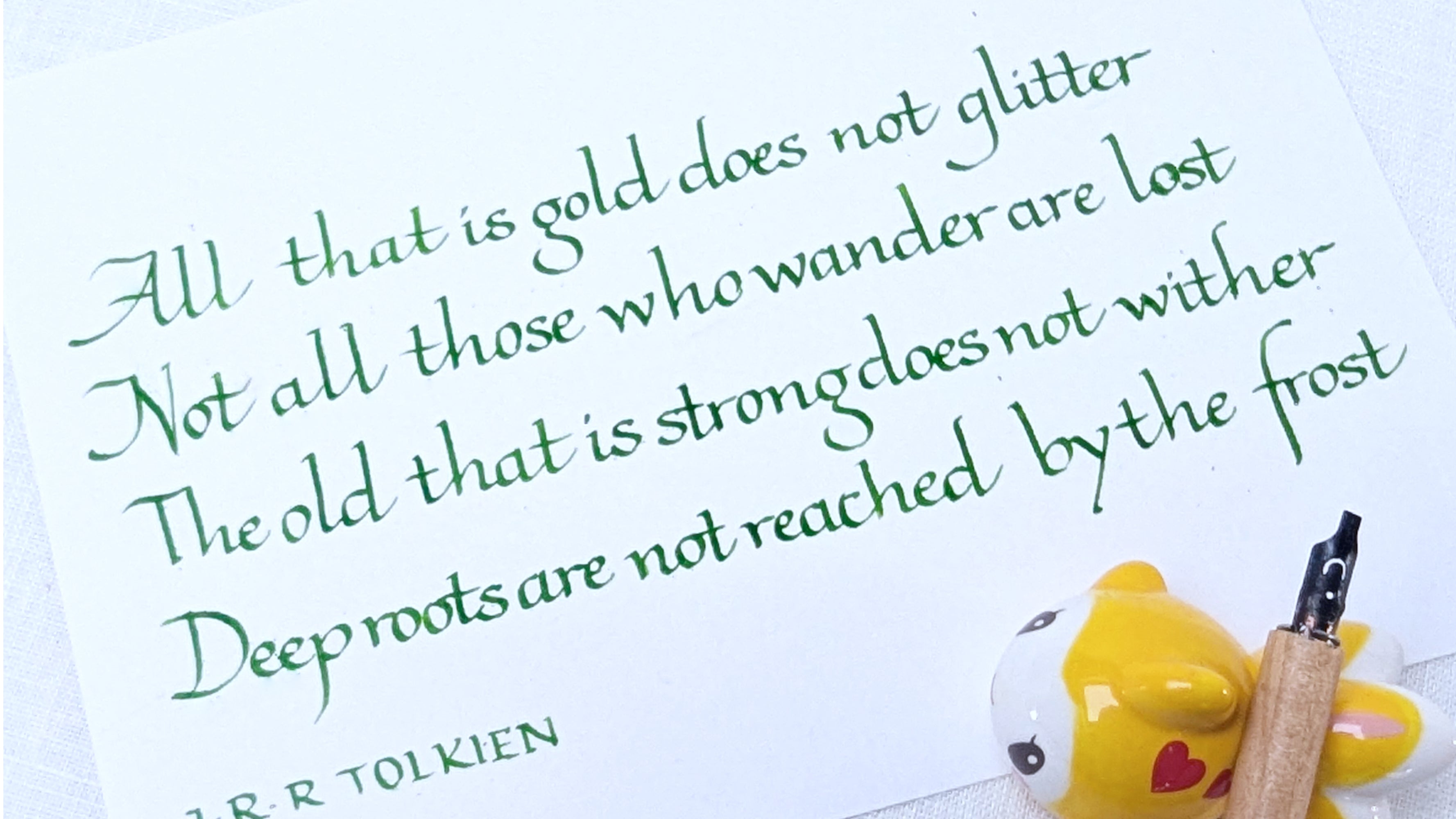 Hand written Calligraphy hand Italic classic exemplar: Quotation from J.R.R. Tolkien on a white background with a yellow mug and pen.