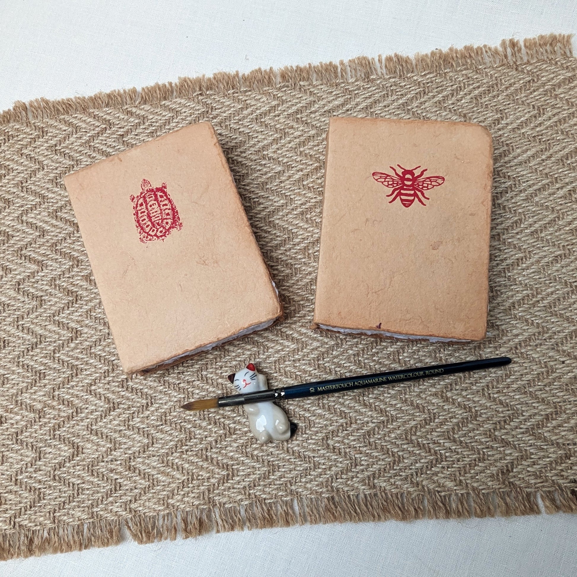 Two small brown books with red embossed designs on a textured surface with a paintbrush and stone.