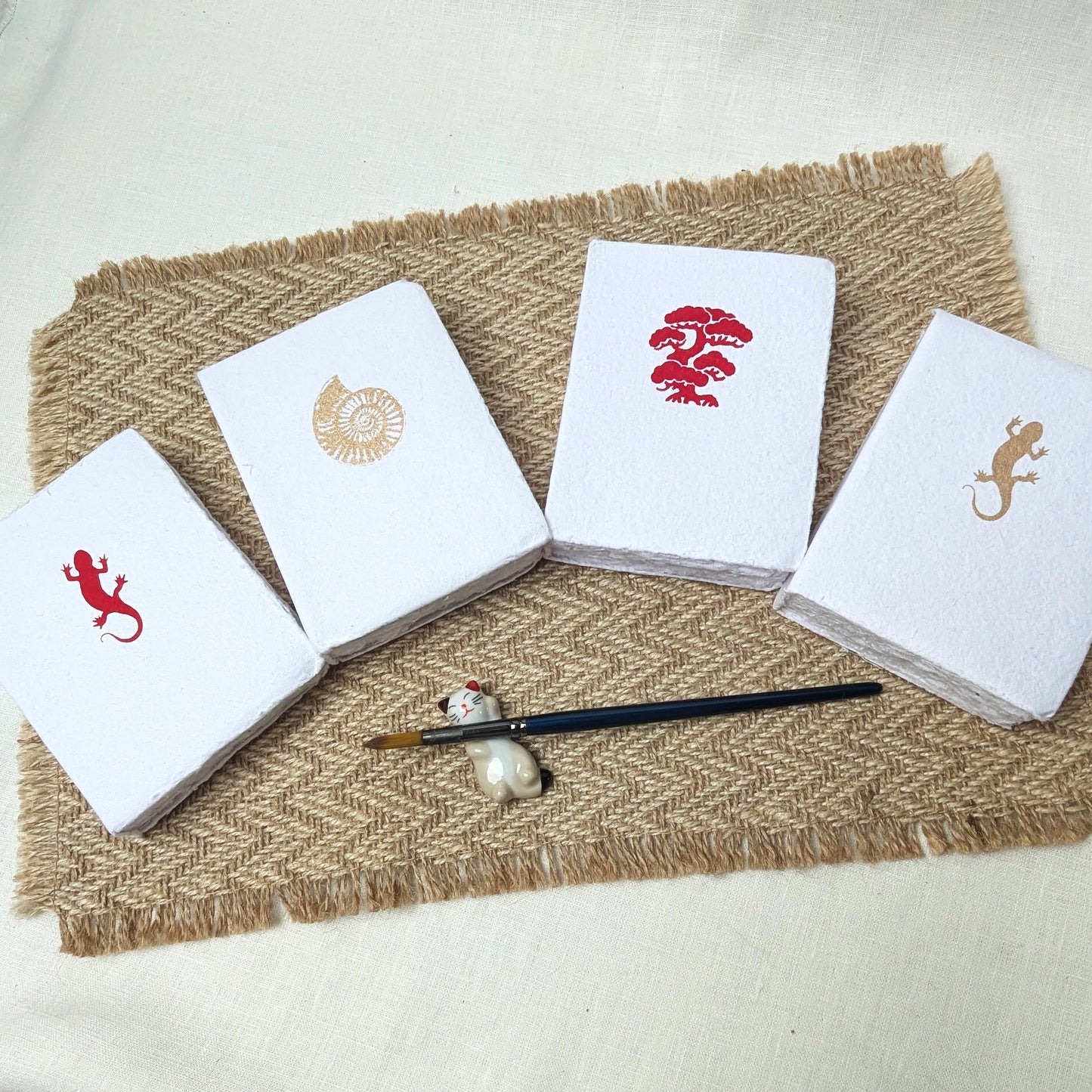 Four white cards with red and gold designs on a textured brown mat with a brush and pearl.