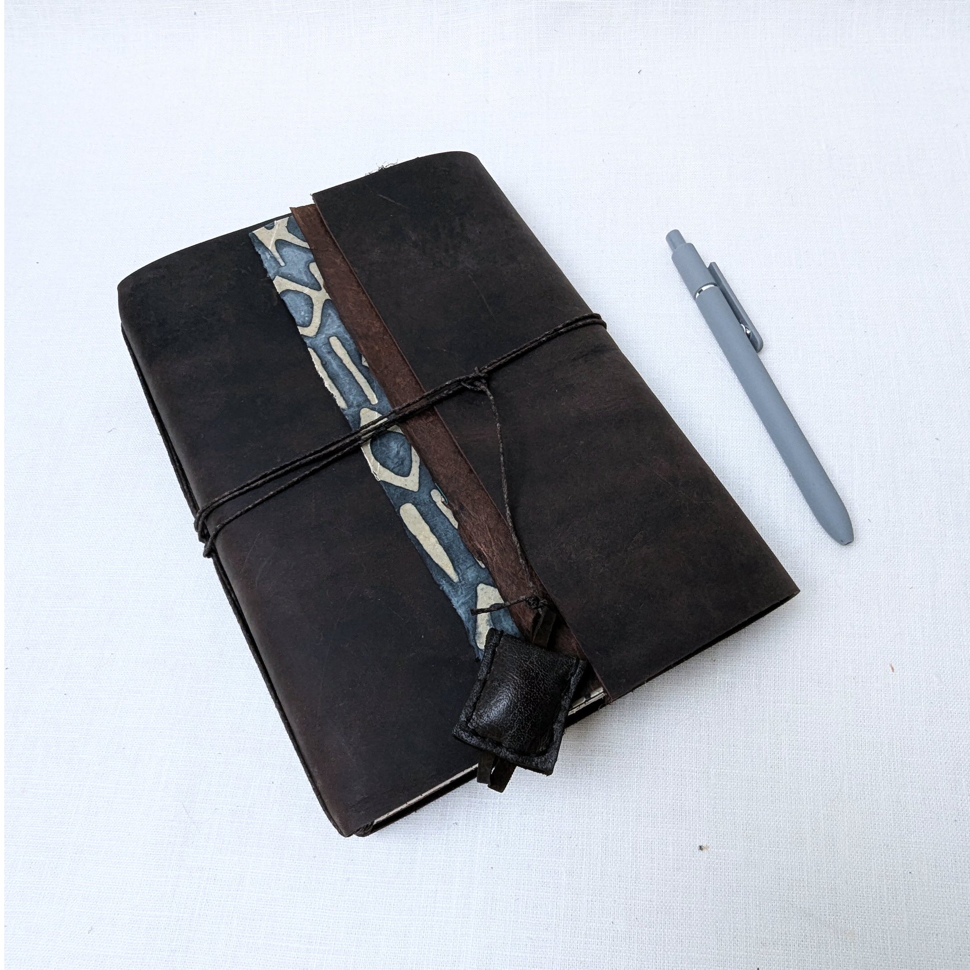 Brown leather notebook with patterned strap and gray pen on a white background