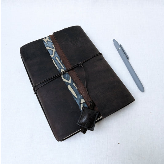 Brown leather notebook with patterned strap and gray pen on a white background