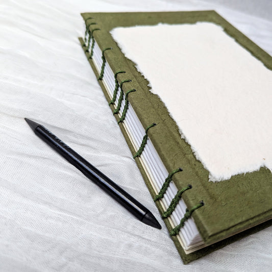 Green sketchbook with white pages and a pen on a white fabric background
